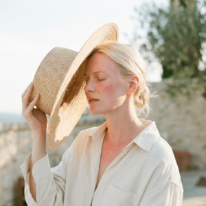 Femme à la peau claire se protégeant le visage du soleil avec un chapeau de paille, légères rougeurs visibles sur la pommette signe d'allergie solaire